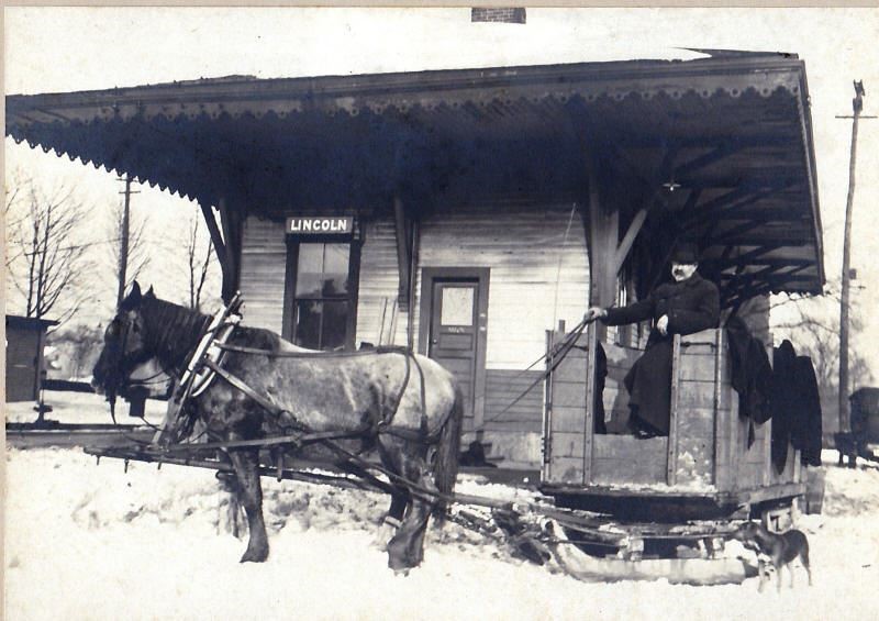 Historic photo of Lincoln train station with horse pulling sled wagon with man standing on it.